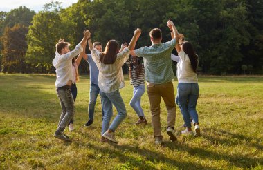 Group of young people having fun and play roundelay dancing in circle in park. Company of funny friends holding hands while walking in circle in park on sunny summer day. Concept of friendship.