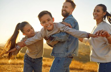 Güneşli bir günde eğlenen ve uçak oynayan dört kişilik mutlu bir aile. Anne, baba ve çocukları gün batımında açık alanda yürüyorlar..