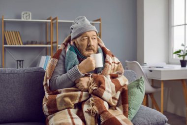Ev tedavisi. Kıdemli adam evdeki kanepede otururken ısınıyor ve kendini sıcak içecekle şımartıyor. Donmuş emekli şapkalı ve battaniyeye sarılı bir şekilde elinde fincanla odada oturuyor..