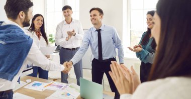 Mutlu gülümseyen şirket çalışanları modern ofiste el sıkışıyorlar. İş adamları, iş arkadaşlarıyla başarılı bir ortaklıktan memnuniyet duyuyorlar ve kapalı bir toplantıda onları alkışlıyorlar..