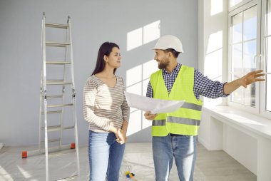 İnşaat işçisi ya da inşaat ustası kağıt planıyla ev sahibiyle konuşuyor. Dost canlısı, kasklı ve üniformalı bir adam yeni evdeki merdivenle odada genç bir bayanla konuşurken konuşuyor.