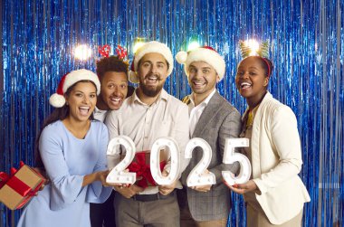 Yeni yıl partisinde, soyut mavi parlak arka planda, elinde Noel 'i kutlayan 2025 numaralı bir yazı bulunan mutlu, çok ırklı arkadaşlar topluluğu. Kış tatili kavramı.