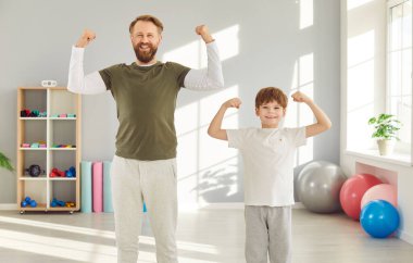 Komik baba ve oğul formda ilerlemelerini gösteriyor ve kaslarını muzaffer pozlarda sergiliyorlar. Parlak spor salonunda birlikte poz veren erkek ve çocuk portresi. Spor ailesi konsepti.