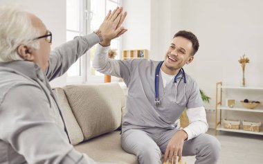 Genç mutlu doktor fizyoterapist, hemşire ya da bakıcı evdeki ya da klinikteki koltukta oturan yaşlı ve gri saçlı bir adama beşlik çakıyor. Emeklilik konsepti.