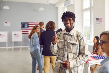 Genç mutlu Afrikalı Amerikalı seçmen oy merkezinin önünde kuyrukta bekliyor ve oy kullanma merkezindeki işaret levhasıyla kameraya neşeli bakıyor. Seçim merkezinde oy pusulası tutan adam.