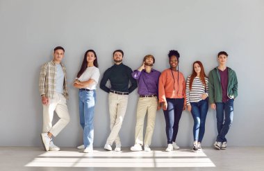 Farklı etnik kökenlere sahip yedi kendine güvenen insan rahat bir duruşla öne bakıyor. Şık bir şekilde gülümseyen genç kadın ve erkekler, arka planda, güneş ışığıyla birlikte duvarın önünde sıralanırlar..