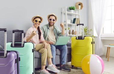 Mutlu gülümseyen çift koltukta bavullarıyla oturuyor ve dizüstü bilgisayarla bilet ayırtıyorlar. Plaj şapkası takan genç bir adam ve kadın tatil gezisine hazırlanıyor. Tatil kavramı.