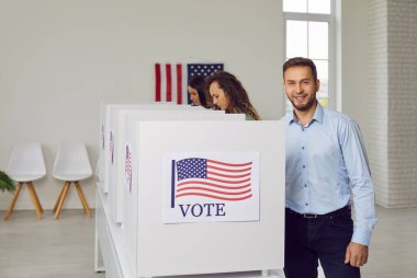 ABD 'de başkanlık seçimlerinde oy kullanan bir grup Amerikalı seçmen. Beyaz oy verme kabininin yanında elinde ABD bayrağı posteriyle duran klasik ofis gömlekli mutlu bir iş adamı.