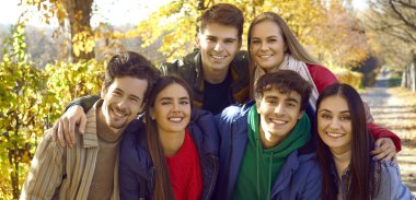 Sonbahar parkında yürüyen ve kucaklaşan bir grup mutlu arkadaşın yakın portresi. Genç, gülümseyen beyaz insanlar dikilip dışarıdaki kameralara bakıyor. Arkadaşlık ve birliktelik kavramı. Pankart.