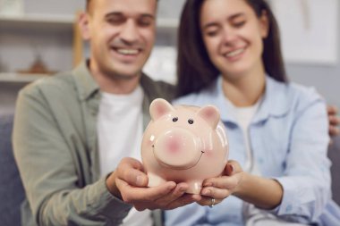 Mutlu genç bir çiftin portresi. Aile bütçelerini planlıyorlar. Kanepede oturuyorlar ve ellerinde domuz kumbarası tutuyorlar. Yatırımlar ve finansal özgürlük kavramı.