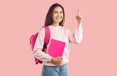 Sırtında sırt çantasıyla pembe stüdyo arka planında izole edilmiş işaret parmağı gösteren mutlu, esmer bir kızın portresi. Eğitim, bilgi kavramı.
