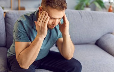 Mutsuz bir adam evdeki oturma odasında bir kanepede oturuyor, üzgün ve stresli görünüyor, baş ağrısından muzdarip. Yorgun ve hasta görünüyor, muhtemelen iş ya da stres yüzünden migren ağrısı çekiyor..