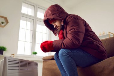 Evdeki kapüşonlu koltukta oturan ve ellerini modern elektrikli ısıtıcıda ısıtmaya çalışan endişeli, genç bir adamın portresi. Isıtma sorunları konsepti.