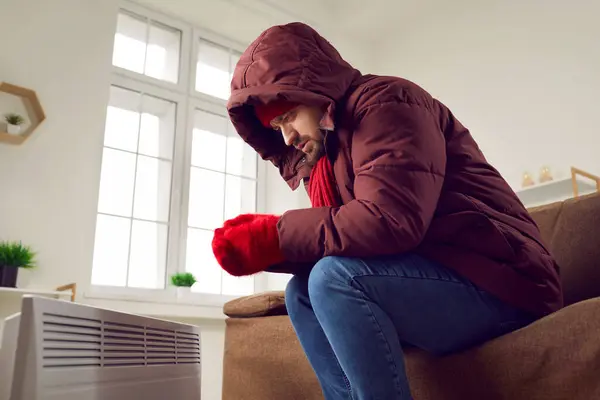stock image Portrait of a young disappointed worried frozen man sitting on sofa in winter outerwear in hood at home and trying to warm his hands on modern electric heater. Heating problems concept.