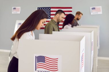 ABD bayrağı taşıyan oy merkezindeki ciddi seçmenler. Bir grup Amerikan vatandaşı gelecekteki başkanlarını seçiyor. Oy verme kabininde duran genç bir kadın adayına oy veriyor. Seçim kavramı