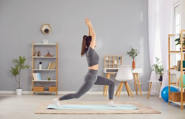 Spor kıyafetleri içinde, evde yoga yapan, oturma odasının iç tarafındaki bir egzersiz minderinin üzerinde duran ve diğer adıyla Ashta Chandrasana Hilal hamlesi yapan güzel bir genç kadının yan görüntüsü.
