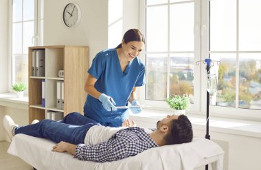 Hastanedeki adamla ilgilenen dost canlısı bir bayan hemşire. Önlüklü mutlu genç bir kadın elinde bir pano, yanında vitamin terapisi serumu, erkek hastayla konuşuyor ve gülümsüyor. 