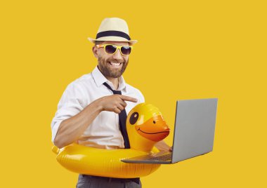 Ofis gömleği, kravat, tatil şapkası, güneş gözlüğü ve komik şişme oyuncak ördek yüzme yüzüğü takan yakışıklı iş adamı yaz tatilinde sarı arka planda tek başına dizüstü bilgisayarda çalışır.