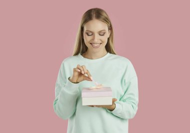 Tatil hediyesi. Neşeli ve meraklı genç bir kadın hediye paketini açarken kurdele çekiyor. Gülümseyen kız doğum günü ya da sevgililer günü hediyesi alıyor, pembe arka planda izole edilmiş..