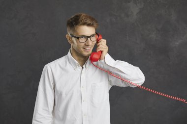 Sabit kırmızı hattan konuşan mutlu bir adamın gri arka planda duran bir stüdyo portresi. Neşeli genç adam telefona cevap veriyor. Gülümseyen müşteri yardım hattını arıyor