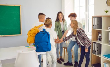 Bir grup mutlu okul çocuğu ve genç, dost canlısı bayan öğretmenleri sınıfta bir çember oluşturup ellerini bir araya getiriyorlar. Okula, eğitime ve takım çalışmasına dönüş konsepti.