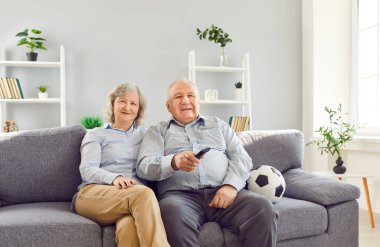 Mutlu, emekli, yaşlı, televizyon kumandası ve futbol topu tutan bir çift portresi. Oturma odasında, spor takımını destekleyen bir kanepede oturuyor..