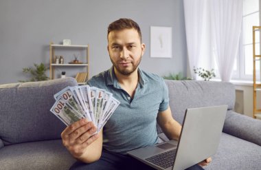 Konsantre olmuş zengin adam evdeki bir kanepede oturmuş, elinde yığınla para tutuyor ve bir dizüstü bilgisayar kullanıyor, parayı kutluyor. Finansal başarı, çevrimiçi çalışma veya kazanma, mutluluk ve refah duygusu.