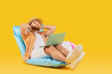 Plajda tatil kıyafeti giyen mutlu bir adam, güneş şapkalı dizüstü bilgisayar, yazın şişme yatakta oturuyor. Boş zaman, eğlence, seyahat, şirket ofisinin dışında çalışmaktan mutlu olmak, serbest çalışmanın tadını çıkarmak.