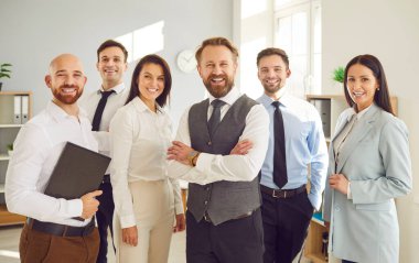 Çok ırklı iş adamları, kameraya bakıp içeride gülümseyen kadın ve erkekler. Şirket çalışanları ya da bir grup personel güvenle ofiste duruyorlar. Takım çalışması konsepti.