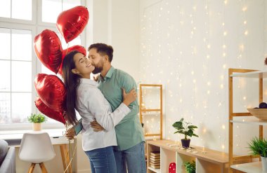 Elinde kırmızı folyo balonlar tutan genç ve mutlu bir adam tatilde güzel bir kadını tebrik ediyor, sarılıyor ve onu öpüyor. Aşık mutlu bir çift. Dikkat, şefkat ve sevgililer günü konsepti.