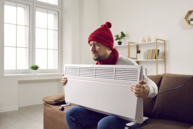 Sıcak giyimli, elinde elektrikli ısıtıcı tutan adam evde üşüyormuş. Örgülü şapka ve eşarp takan üzgün genç adam merkezi ısıtmada sorun yaşıyor.