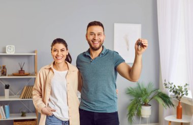 Yeni evlerinde ya da dairelerinde mutlu ve sevgi dolu bir çift portresi. İki neşeli, neşeli genç ev sahibi eğleniyor, oturma odasında duruyor, anahtarı tutuyor ve gülümsüyor. İpotek kavramı