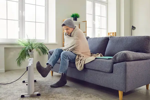stock image Side view of a young frozen man sitting on sofa in the living room at home wrapped in blanket and trying to warm his feet putting his legs on electric heater. Heating problems concept.