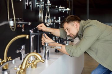 Tesisatçıda çalışan bir adam, farklı muslukları ve muslukları karşılaştırarak evdeki banyo yenilemesine hazırlanıyor. Tesisat ve tasarım için her seçeneği değerlendiriyor..
