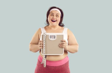 Stüdyonun mavi arka planında spor giysileri giymiş, elinde terazi tutan ve elinde bant tutan mutlu ve heyecanlı şişman bir kadının portresi. Obezite, ağırlık kontrolü, fitness ve spor konsepti.