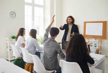 Lise öğrencileri ders sırasında sınıfın masalarında oturuyorlar. Erkek öğrenci, genç ve dost canlısı bir öğretmene cevap vermek için elini kaldırıyor. Okula dönmek, eğitim kavramı..