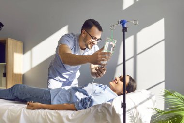 Dost canlısı erkek hemşire, vitamin tedavisi gören mutlu kadın hastaya serum veriyor. Açık mavi önlüklü gülümseyen adam, tıp bürosunda yatmakta olan neşeli genç kadın için damar içi çizgiler çiziyor. 