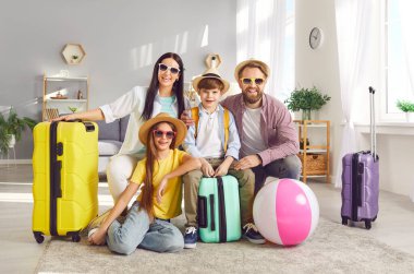 Çocukları olan mutlu ebeveynlerin portresi, bavullarıyla yerde oturan ve tropikal tatil köyüne uçmaya hazır kameralara bakan kız ve oğul. Aile tatili ve tatil gezisi.