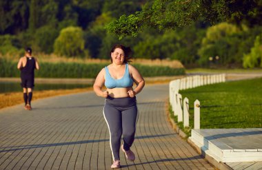 Genç şişman kadın yazın dışarıda koşu antrenmanı yapıyor. Güzel, güneşli bir sabahta yeşil bir parkta asfalt bir yolda koşan şişman, mutlu bir kız. Fitness konsepti