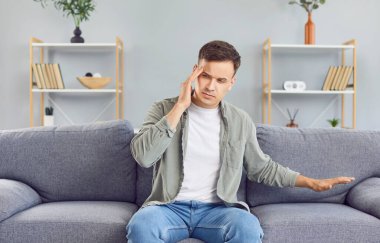 Üzgün ve yorgun bir adam evdeki bir kanepede oturuyor, baş ağrısı ya da migrenden muzdarip. Stres ve acı yüzünden belli oluyor. Rahatsızlığı ve uğraştığı sorunu vurguluyor..