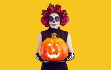 Cadılar Bayramı 'nı ya da Ölüler Günü' nü kutluyoruz. Meksika tarzı yüz sanatı olan kadın stüdyoda turuncu arka planda parlayan balkabağı pozu veriyor. Parlak makyajlı ve şakayık çelenkli bir kadın..