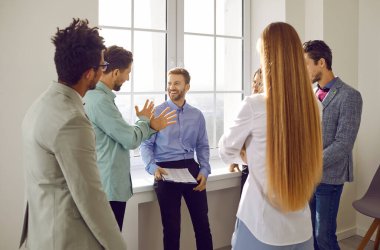 İş ekibi ofiste bir iş toplantısı sırasında tartışıyor. Bir grup genç insan ofis penceresinin önünde duruyor, konuşuyor, fikir alışverişinde bulunuyor ve ilginç önerilerde bulunuyorlar.