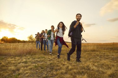 Bir grup genç turist sırt çantalarıyla sahada yürüyorlar. Mutlu insanlar, erkek ve kadınlar gün batımında doğa yürüyüşüne çıkıyorlar. Macera, seyahat ve turizm konsepti.
