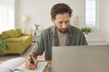 Ciddi odaklı sakallı yetişkin bir adam evde oturmuş, dizüstü bilgisayar kullanıyor, internetten bilgi okuyor, web sitesi izliyor, deftere yazı yazıyor. Çevrimiçi eğitim kavramı