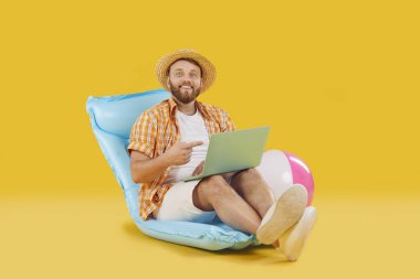 Sarı arka plandaki neşeli adam dizüstü bilgisayarlı şişme yatakta oturuyor. Şişme yatak manzarası tatil heyecanı şişirilebilir yatak boş zamanlarında yaklaşan tatil beklentisi