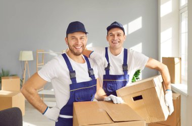 Nakliye ve dağıtım şirketinde gülümseyen iki profesyonel erkek işçinin portresi. Tulumlu, karton kutular taşıyan ve kameraya gülümseyen yakışıklı beyaz adamlar. Taşınma günü konsepti.