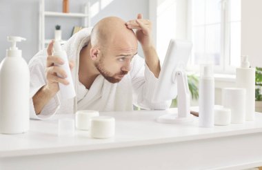 Endişeli, beyaz bornozlu genç adam. Hasarlı kökler ve erken saç dökülmesi tedavisi için şampuanlar ve spreylerle masada oturuyor. Aynaya bakıyor, üst ve alnını ovuyor.