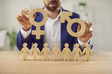 İş adamı, erkek ve kadının ahşap sembollerini insan figürleri zincirinin üstünde tutuyor. Bir işe başvururken cinsiyetler arasındaki fark. Cinsiyet eşitliği ve eşitlik kavramı.