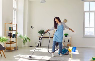 Gülümseyen mutlu genç kadın elektrikli süpürge halısıyla temizlik yapıyor ve evdeki oturma odasında dans ediyor. Yerleri süpüren kadın hademe. Temizlik, temizlik, ev işleri ve ev işleri.