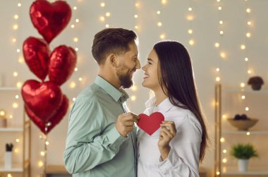 Genç mutlu esmer kadın ve kağıt kalp tutan adam kırmızı folyo balonlar ve arka planda ışıklarla birbirlerine bakıyorlar. Aşık mutlu bir çift. Sevgililer Günü konsepti.